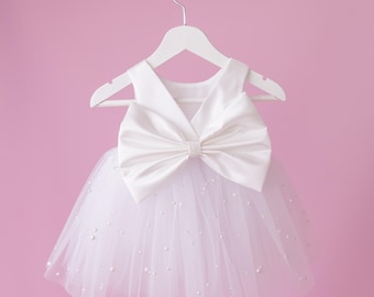 May include: A white baby dress with a large bow and a tulle skirt adorned with small pearls. The dress is hanging on a white hanger against a pink background. The dress is likely for a special occasion.