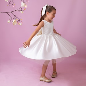 May include: A young person twirls in a white satin dress with a full skirt and a bow in the back. The dress is paired with gold-colored shoes. A white bow is in their hair. The background is a solid pink color.