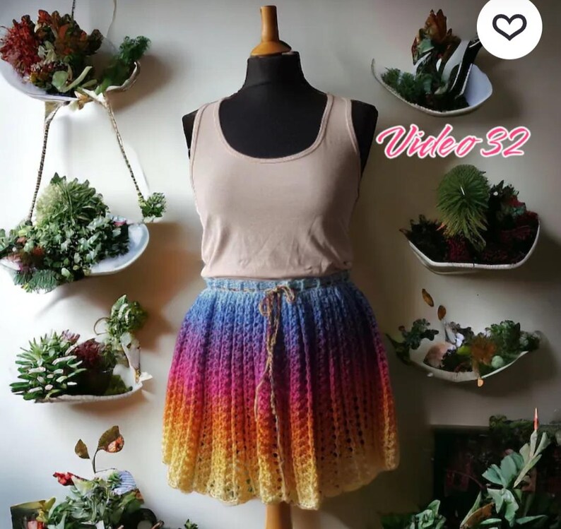 Puede incluir: Una camiseta sin mangas beige y una falda de ganchillo arco&iacute;ris en un maniqu&iacute;. La falda hace la transici&oacute;n de azul a morado, naranja y amarillo. El texto "Video 32" es visible. El fondo presenta jardineras colgantes con suculentas.