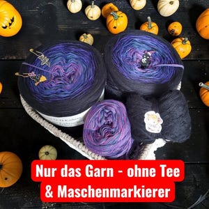 Könnte beinhalten: Drei Garnknäuel mit einem Farbverlauf in Schwarz, Lila und Blau. Zwei Knäuel sind groß und rund, eines ist kleiner. Das Garn enthält Maschenmarkierer aus Metall in Form einer Fledermaus und einer Katze. Ein Kuh-Aufkleber ist auf dem kleinen Knäuel.