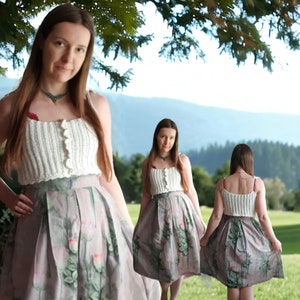 Puede incluir: Un top de ganchillo color crema con botones en la parte delantera, combinado con una falda plisada con estampado floral en tonos rosas y verdes. El conjunto se lleva al aire libre con un fondo natural.