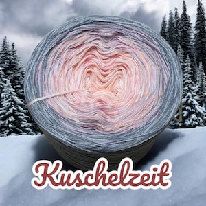 Könnte beinhalten: Eine Garnrolle mit einem Farbverlauf von rosa, orange und grau. Das Garn ist zu einer Kugel gewickelt und hat ein Etikett mit der Aufschrift "Kuschelzeit".
