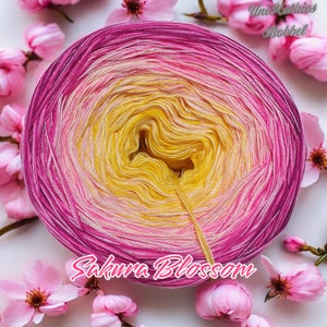 Könnte beinhalten: Ein lebendiger Garnball mit Farbverlauf von tiefem Pink zu Gelb. Rosa Kirschblüten umgeben das Garn, mit dem Text "Sakura Blossom" in Schreibschrift unten. Perfekt zum Stricken und Häkeln.