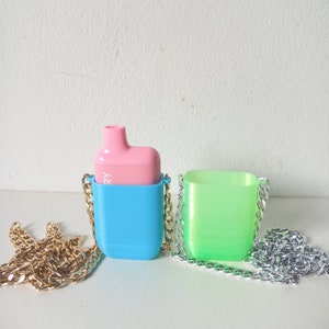 Op de afbeelding: Twee kleine, kleurrijke hoesjes met kettingbandjes. Het hoesje links is lichtblauw met een roze bovenkant en een gouden ketting. Het hoesje rechts is groen met een zilveren ketting.