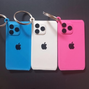 Könnte beinhalten: Drei rechteckige Schlüsselanhänger in Blau, Weiß und Pink, die wie ein Smartphone gestaltet sind. Jeder Anhänger hat ein schwarzes Apfel-Logo in der Mitte, ein Kamera-Modul mit drei Linsen in der Ecke und einen silbernen Metallring zur Befestigung.