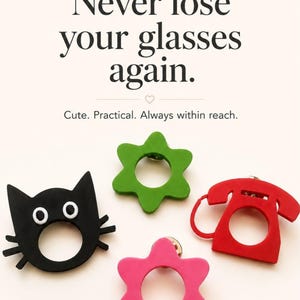 Könnte beinhalten: Vier Brillenhalter in Form einer schwarzen Katze, eines roten Telefons, einer grünen Blume und einer pinken Blume. Der Text lautet: Never lose your glasses again. Cute. Practical. Always within reach. Diese Artikel dienen als Anstecknadeln.