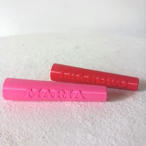 May include: Two cylindrical objects, one pink and one red, each with a name embossed on the side. The pink object reads "MARIA", while the red object reads "LEANDRO". Both are set against a white background.