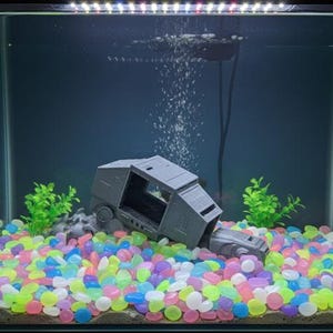 Puede incluir: Un modelo gris de AT-AT sumergido en un acuario, rodeado de guijarros coloridos y brillantes. Plantas de plástico verde se colocan a ambos lados del modelo. Las burbujas se elevan desde una piedra de aire, creando una escena submarina.