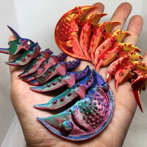 May include: A set of 3D printed trilobites in various colors, including red, orange, green, blue, and purple. The trilobites are arranged in a semi-circle on a hand.