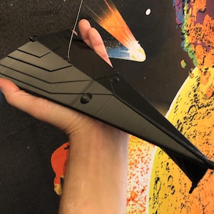May include: A black plastic model of a spaceship, held in a person's hand. The model has a sleek, angular design with a pointed nose and a flat bottom. The background is a colourful, abstract design.