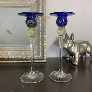 May include: A pair of glass candle holders with blue and yellow glass accents. The candle holders have a wide base and a tall, slender stem.
