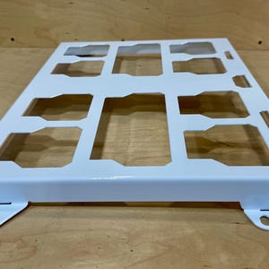 May include: A white metal grid with rectangular and trapezoidal cutouts. The grid has a rectangular shape with a raised border and mounting tabs on the corners. The object is likely a component or part of a larger structure.