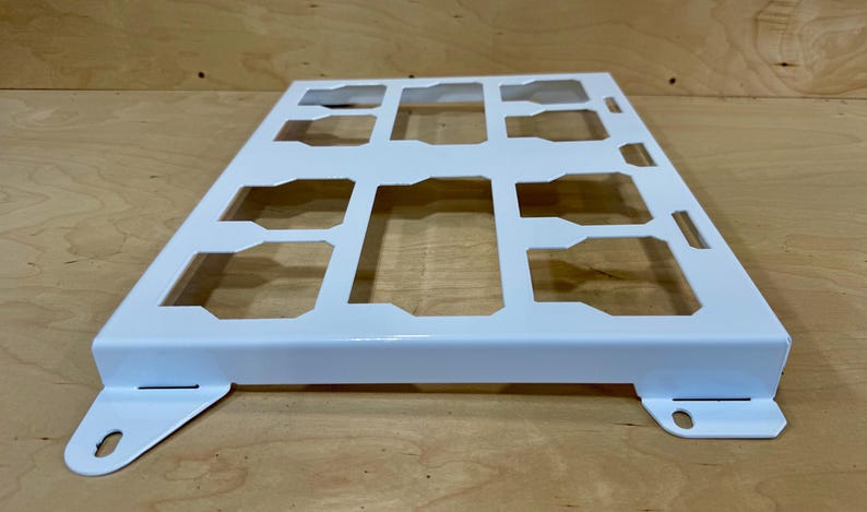May include: A white metal grid with rectangular and trapezoidal cutouts. The grid has a rectangular shape with a raised border and mounting tabs on the corners. The object is likely a component or part of a larger structure.