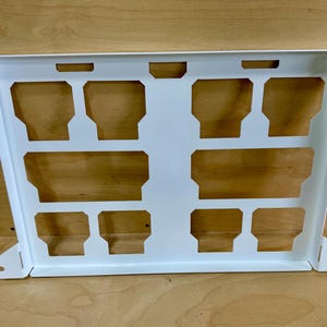 May include: A white metal bracket with a series of rectangular cutouts. The bracket has a rectangular shape with angled sides and mounting holes on the corners. The cutouts are arranged in a grid pattern.