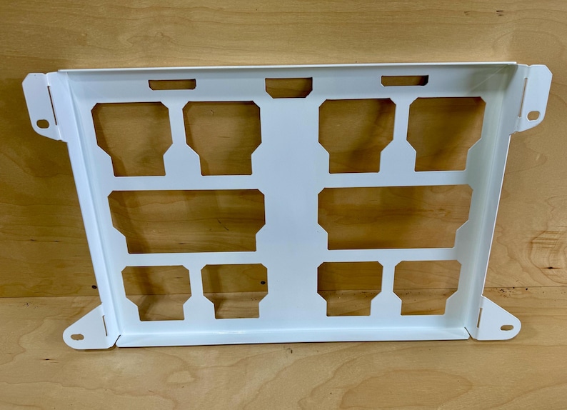 May include: A white metal bracket with a series of rectangular cutouts. The bracket has a rectangular shape with angled sides and mounting holes on the corners. The cutouts are arranged in a grid pattern.