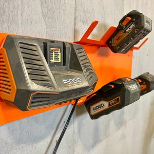 Puede incluir: Un soporte de metal naranja montado en la pared con tres bater&iacute;as de herramientas el&eacute;ctricas Ridgid. El soporte tiene un cable de alimentaci&oacute;n negro y una etiqueta de advertencia. Las bater&iacute;as son negras y naranjas con el logotipo de Ridgid y 18V impreso en ellas.
