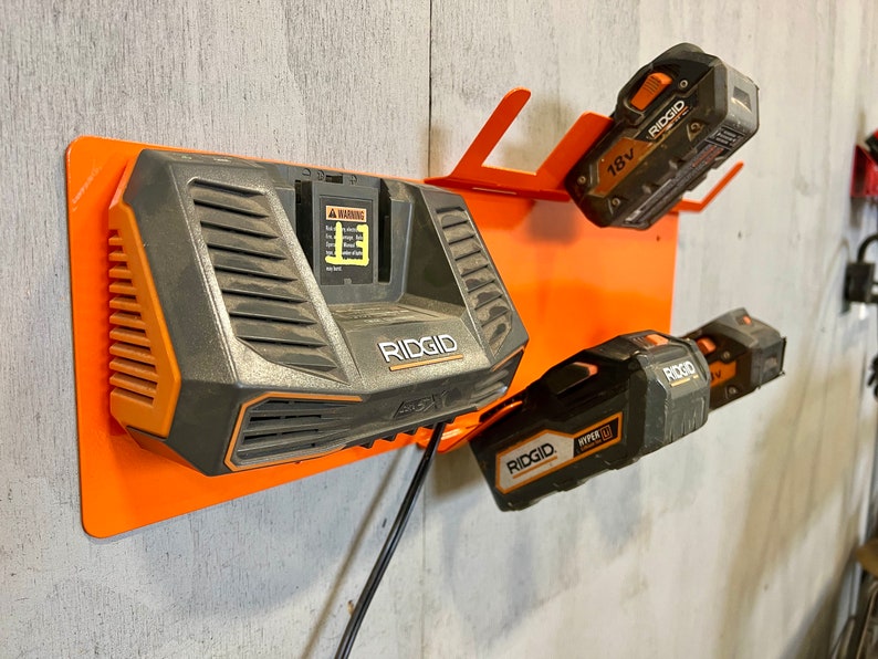 Puede incluir: Un soporte de metal naranja montado en la pared con tres bater&iacute;as de herramientas el&eacute;ctricas Ridgid. El soporte tiene un cable de alimentaci&oacute;n negro y una etiqueta de advertencia. Las bater&iacute;as son negras y naranjas con el logotipo de Ridgid y 18V impreso en ellas.