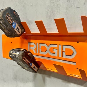 Puede incluir: Soporte de pared de metal naranja con la palabra "RIDGID" recortada en el centro. El soporte tiene seis ranuras para sujetar bater&iacute;as. Dos bater&iacute;as negras y naranjas est&aacute;n en las ranuras. Las bater&iacute;as est&aacute;n etiquetadas como "OCTANE 9.0 Ah" y "OCTANE 3.0 Ah".