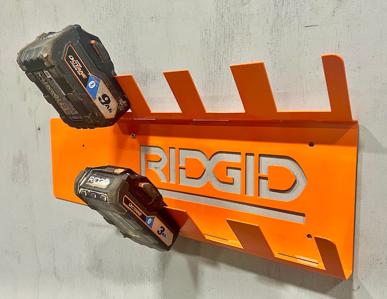 Puede incluir: Soporte de pared de metal naranja con la palabra "RIDGID" recortada en el centro. El soporte tiene seis ranuras para sujetar bater&iacute;as. Dos bater&iacute;as negras y naranjas est&aacute;n en las ranuras. Las bater&iacute;as est&aacute;n etiquetadas como "OCTANE 9.0 Ah" y "OCTANE 3.0 Ah".