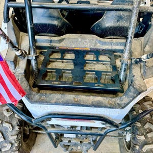 May include: A black metal cargo rack is mounted on the back of a white and gray Polaris ATV. The ATV has large, rugged tires and a black roll cage. An American flag is attached to the side.