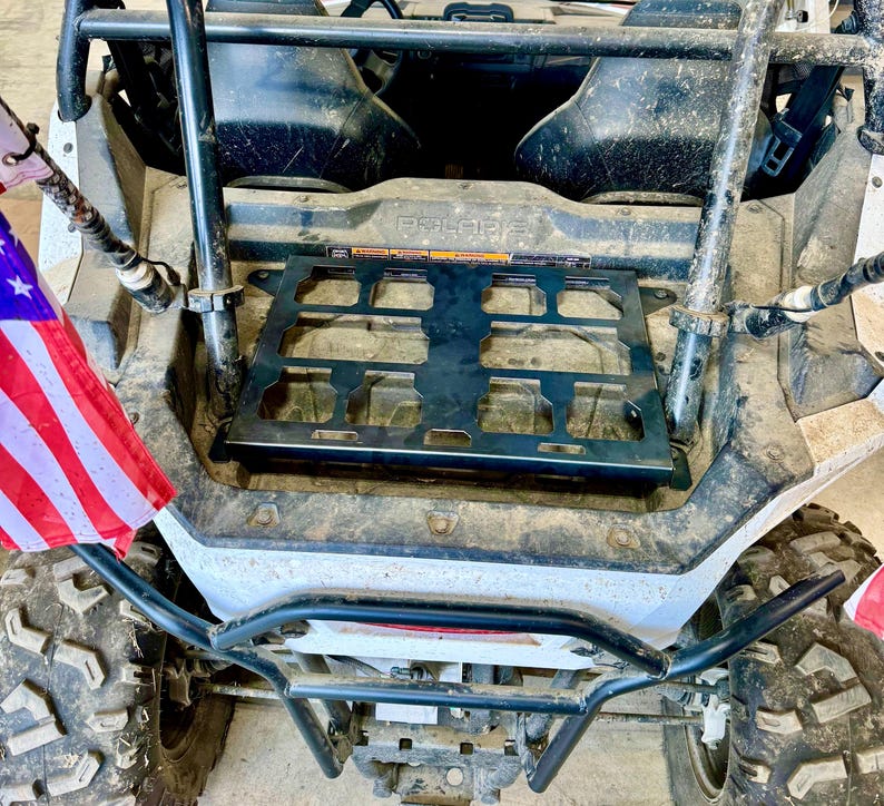 May include: A black metal cargo rack is mounted on the back of a white and gray Polaris ATV. The ATV has large, rugged tires and a black roll cage. An American flag is attached to the side.