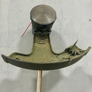 May include: A close-up of a weed trimmer head with a black plastic housing and a silver metal shaft. The head is covered in green grass clippings.