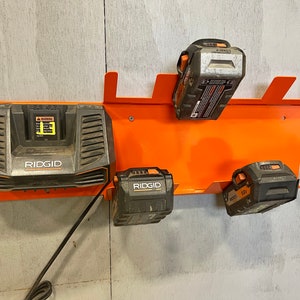 Puede incluir: Un estante de almacenamiento de bater&iacute;as de pared naranja con cuatro bater&iacute;as negras Ridgid, incluido un cargador con un cable de alimentaci&oacute;n negro.