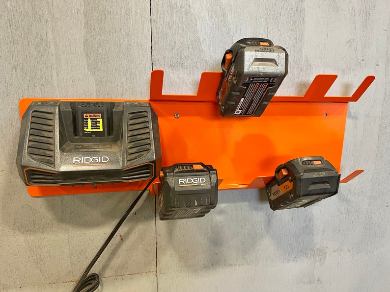 Puede incluir: Un estante de almacenamiento de bater&iacute;as de pared naranja con cuatro bater&iacute;as negras Ridgid, incluido un cargador con un cable de alimentaci&oacute;n negro.