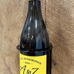May include: A bottle of A to Z Wineworks 2021 Oregon Pinot Gris wine in a black metal bottle holder against a wood background.
