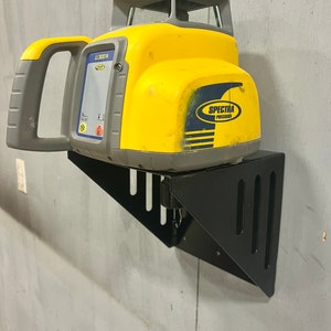 May include: A yellow and gray Spectra Precision laser level mounted on a black metal wall mount. The laser level has a handle and a clear window on the top.