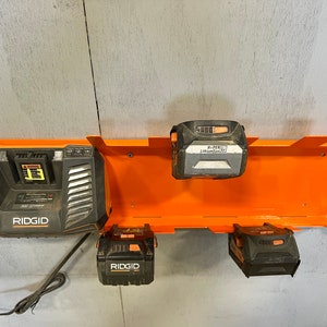Puede incluir: Un soporte de pared naranja para almacenamiento de bater&iacute;as con cuatro bater&iacute;as. El soporte tiene ranuras para sujetar las bater&iacute;as. Las bater&iacute;as son negras y naranjas y tienen la marca "RIDGID" impresa en ellas. Una de las bater&iacute;as es un cargador.