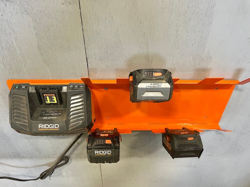 Puede incluir: Un soporte de pared naranja para almacenamiento de bater&iacute;as con cuatro bater&iacute;as. El soporte tiene ranuras para sujetar las bater&iacute;as. Las bater&iacute;as son negras y naranjas y tienen la marca "RIDGID" impresa en ellas. Una de las bater&iacute;as es un cargador.
