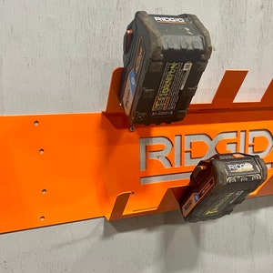 Puede incluir: Soporte de pared de metal naranja con la palabra "RIDGID" recortada, dise&ntilde;ado para sujetar dos bater&iacute;as de herramientas el&eacute;ctricas negras y amarillas.