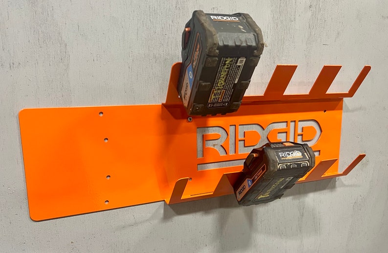 Puede incluir: Soporte de pared de metal naranja con la palabra "RIDGID" recortada, dise&ntilde;ado para sujetar dos bater&iacute;as de herramientas el&eacute;ctricas negras y amarillas.