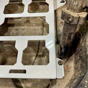 May include: A white metal component with a rectangular frame and multiple cut-out sections. The component is attached to a dark surface with a metal tube and a bolt. The image is taken from an overhead perspective.