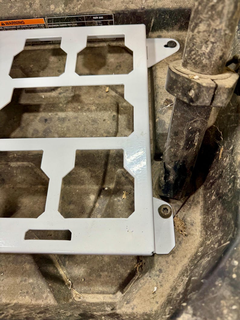 May include: A white metal component with a rectangular frame and multiple cut-out sections. The component is attached to a dark surface with a metal tube and a bolt. The image is taken from an overhead perspective.