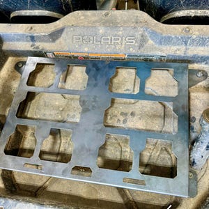 May include: A silver metal accessory with multiple cut-out shapes, likely for a vehicle. The accessory is mounted on a dirty, textured surface, with the brand name "POLARIS" visible. The design suggests it's a functional part.