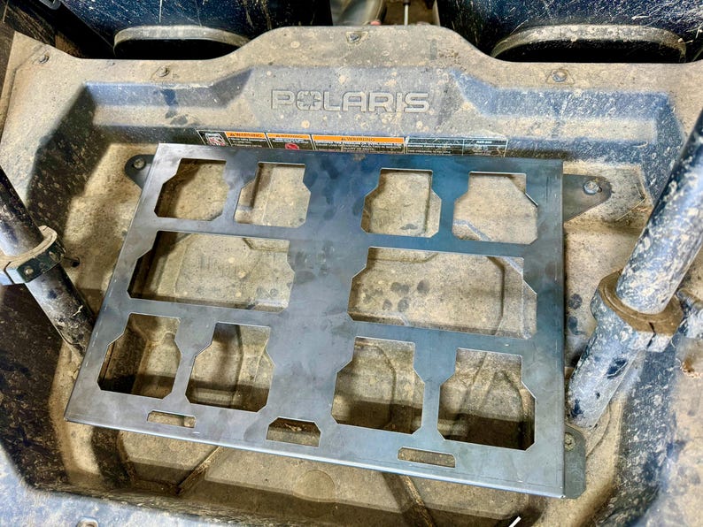 May include: A silver metal accessory with multiple cut-out shapes, likely for a vehicle. The accessory is mounted on a dirty, textured surface, with the brand name "POLARIS" visible. The design suggests it's a functional part.