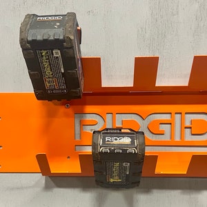 Puede incluir: Soporte de pared de metal naranja con logotipo RIDGID recortado, dise&ntilde;ado para sostener dos bater&iacute;as RIDGID negras.