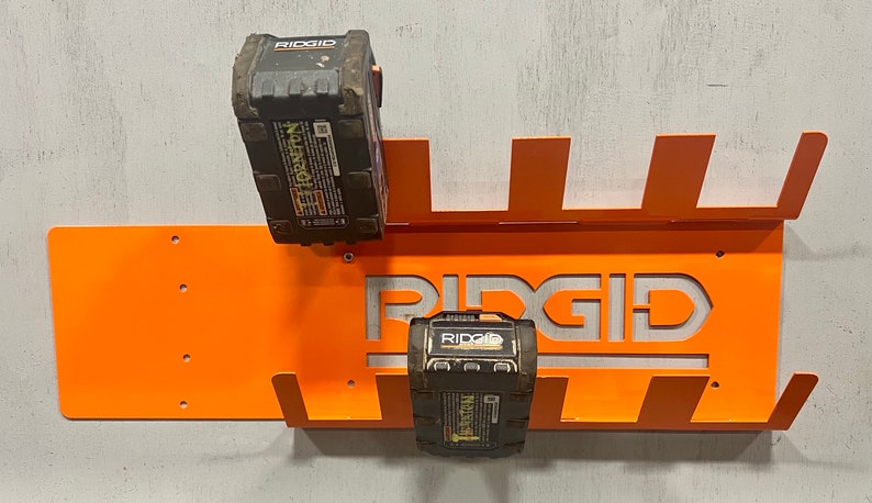 Puede incluir: Soporte de pared de metal naranja con logotipo RIDGID recortado, dise&ntilde;ado para sostener dos bater&iacute;as RIDGID negras.