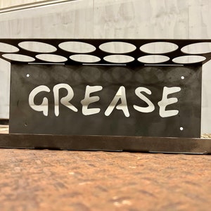 Peut inclure: Un porte-graisse en m&eacute;tal noir avec 12 trous ronds et le mot "GREASE" d&eacute;coup&eacute; en blanc.