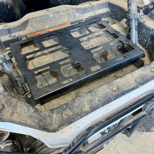 May include: A black metal cargo rack installed in a Polaris off-road vehicle. The rack has a rectangular shape with multiple cut-out sections. Warning labels are visible on the rack. The vehicle's interior is dirty.