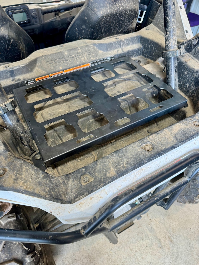 May include: A black metal cargo rack installed in a Polaris off-road vehicle. The rack has a rectangular shape with multiple cut-out sections. Warning labels are visible on the rack. The vehicle's interior is dirty.