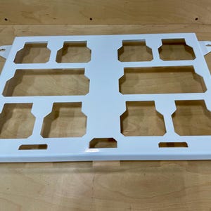 May include: A white metal tray with a rectangular shape and cut-out sections. The tray has a series of rectangular and square openings, with small tabs on the sides. The tray is designed to hold multiple items.