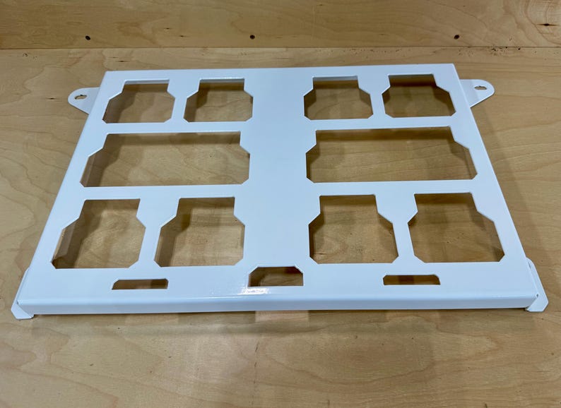 May include: A white metal tray with a rectangular shape and cut-out sections. The tray has a series of rectangular and square openings, with small tabs on the sides. The tray is designed to hold multiple items.