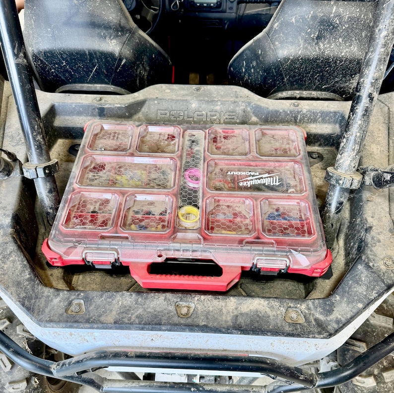 May include: A red and clear Milwaukee Packout organizer with multiple compartments, filled with various small parts. The organizer is placed in a Polaris vehicle, with the brand name visible.