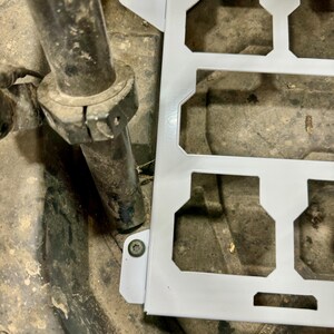 May include: A white metal bracket with multiple rectangular cutouts and mounting tabs. The bracket is positioned near a dark, cylindrical metal component and a textured surface.