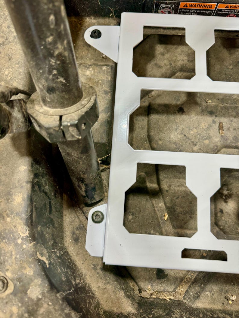 May include: A white metal bracket with multiple rectangular cutouts and mounting tabs. The bracket is positioned near a dark, cylindrical metal component and a textured surface.