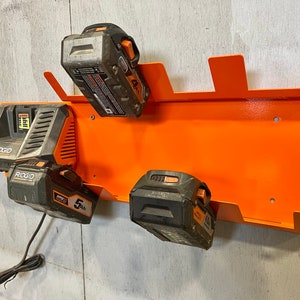 Puede incluir: Soporte de pared de metal naranja con cinco ranuras para guardar bater&iacute;as de herramientas el&eacute;ctricas. Tres bater&iacute;as est&aacute;n en las ranuras, dos son negras y una es gris. Las bater&iacute;as est&aacute;n etiquetadas como "RIDGID" y "5 Ah".