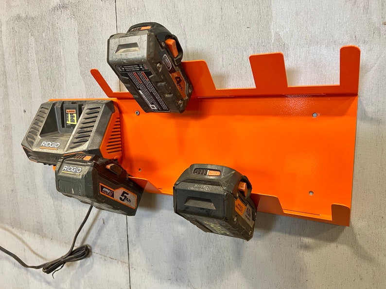 Puede incluir: Soporte de pared de metal naranja con cinco ranuras para guardar bater&iacute;as de herramientas el&eacute;ctricas. Tres bater&iacute;as est&aacute;n en las ranuras, dos son negras y una es gris. Las bater&iacute;as est&aacute;n etiquetadas como "RIDGID" y "5 Ah".
